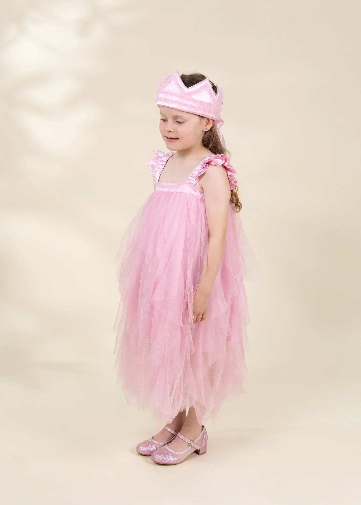 A young girl wearing a light pink tulle dress with ruffled straps, a matching pink headband, and pink shoes, standing against a plain beige studio background.