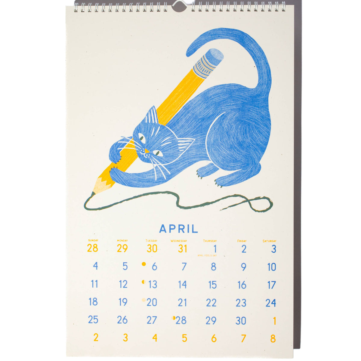 A spiral-bound desk calendar page featuring a blue cat drawing with a yellow pencil on an off-white background, with the word APRIL and a numbered calendar grid below.