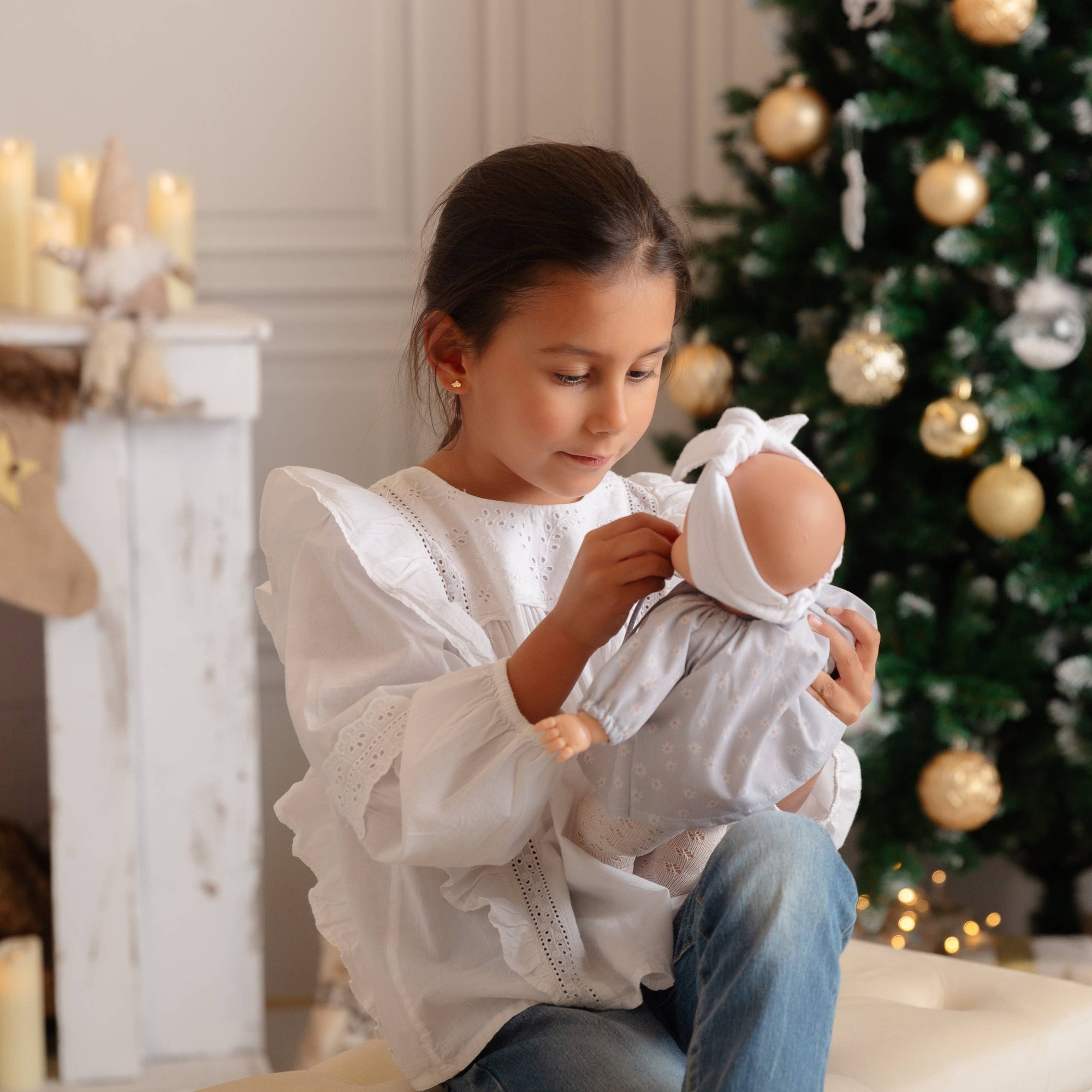 Young girl holding a baby doll in front of a decorated Christmas tree.