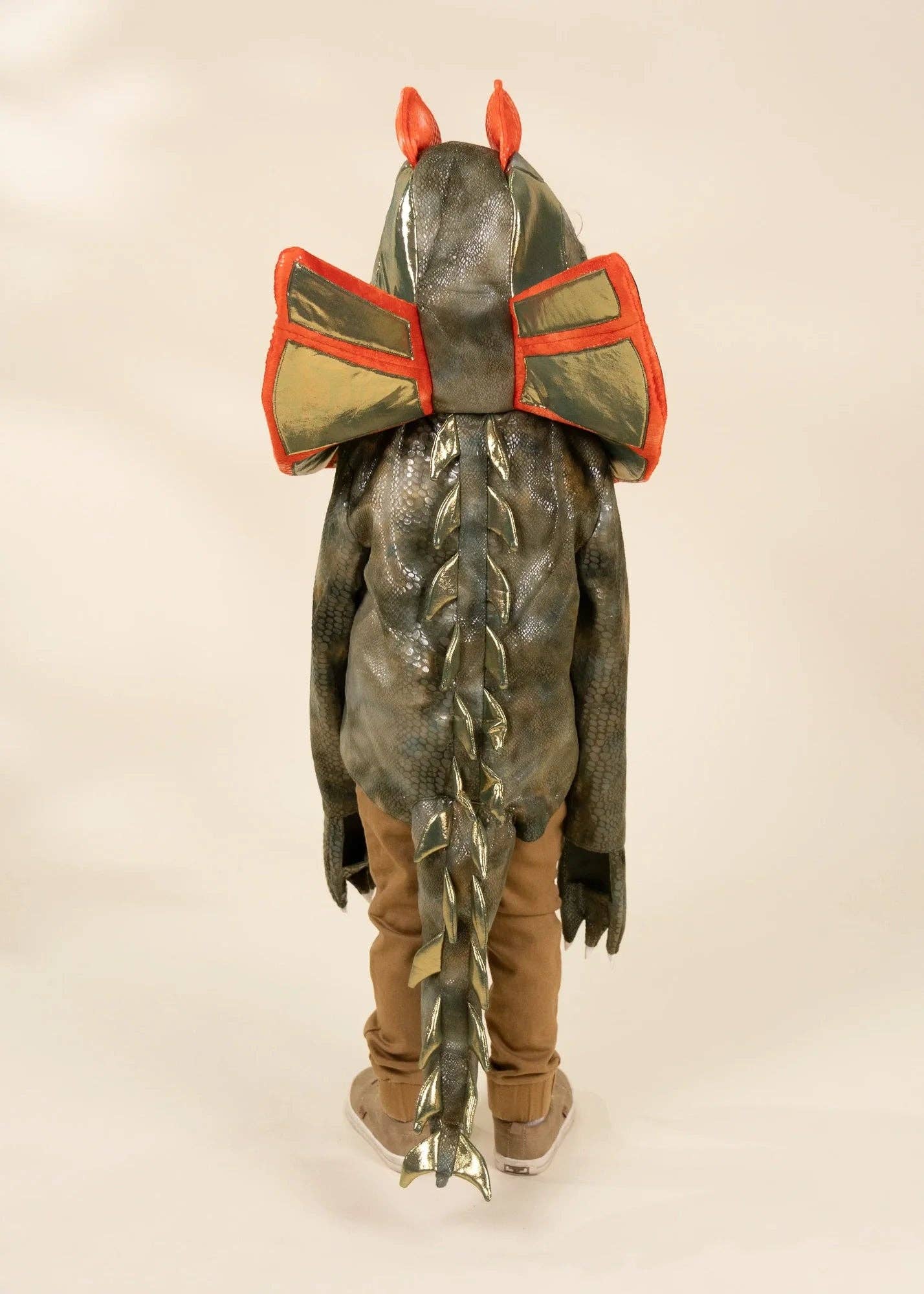 A child viewed from behind wearing a detailed monster or dragon costume, featuring a textured dark gray hood with red horns, metallic gold and red shoulder pieces, a long braided tail, and brown pants, set against a plain beige studio background.