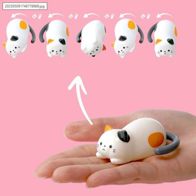 Small white cat-shaped toy with orange spots and gray tail held in a hand against a pink background.