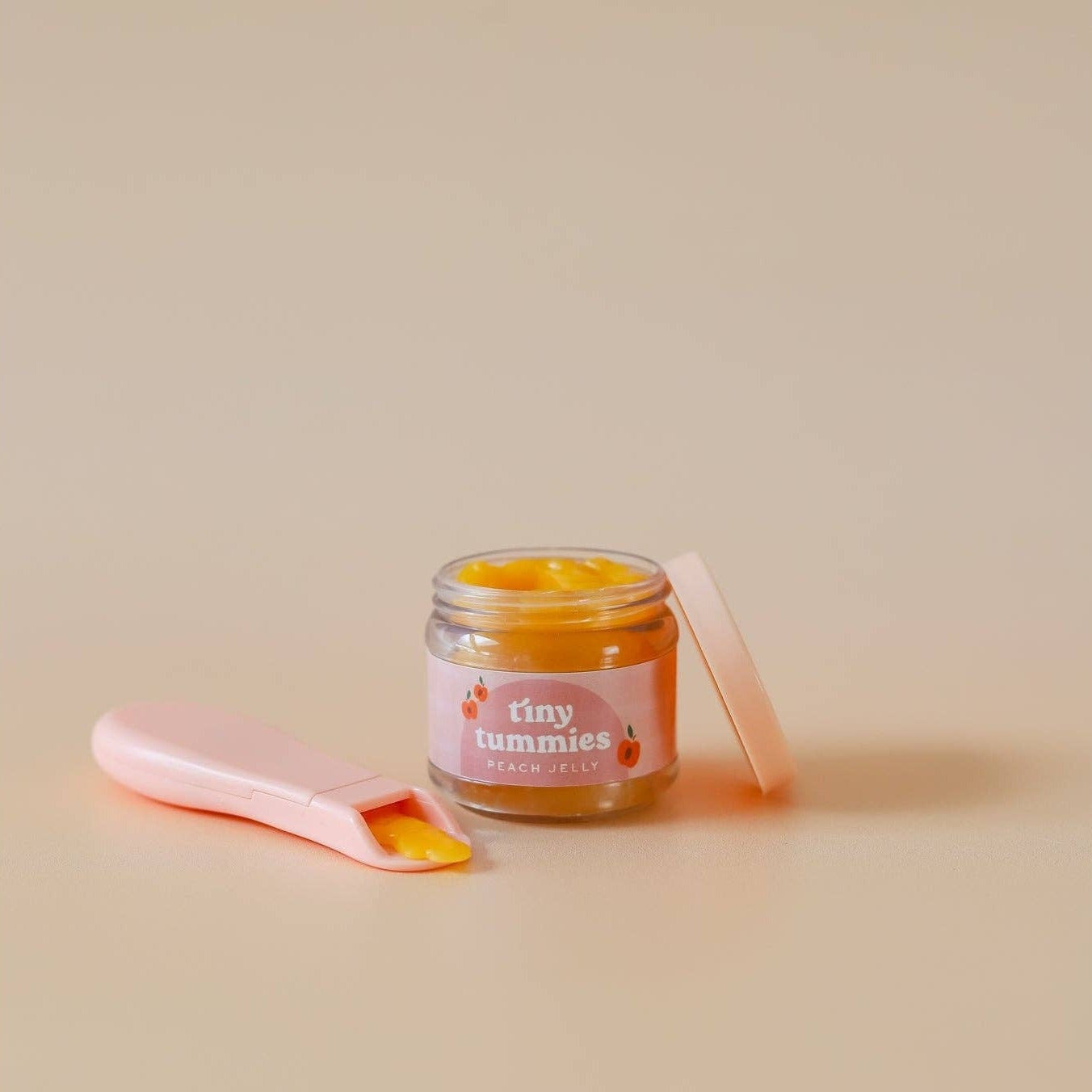 Jar of 'tiny tummies' baby food with a pink spoon on a beige background