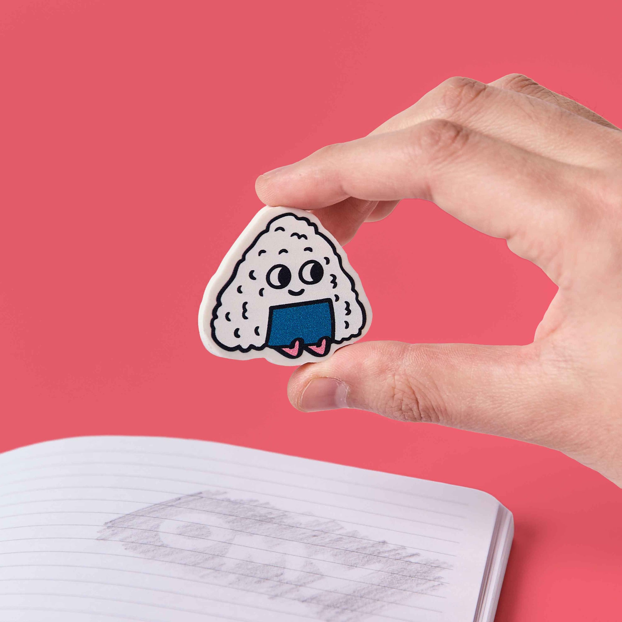 Hand holding a cartoon rice ball sticker over an open notebook with a pink background