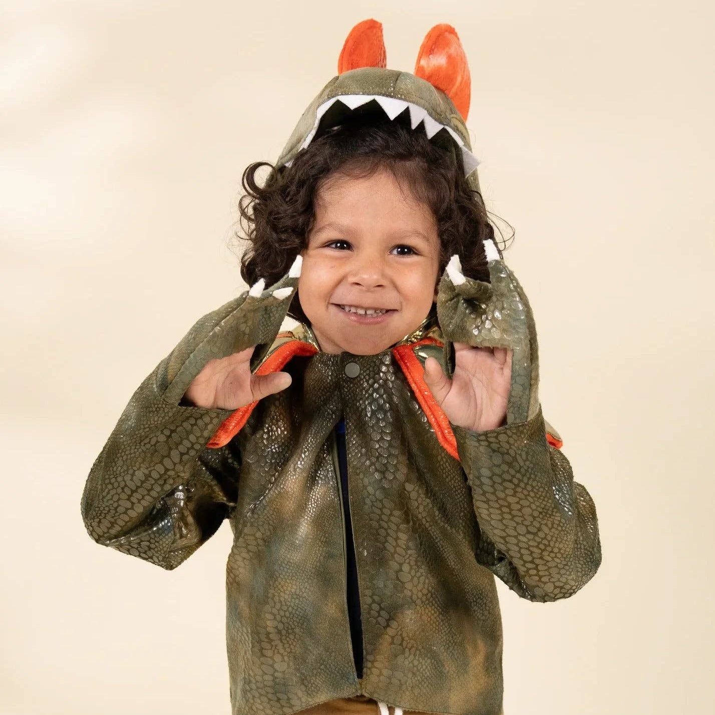 A young child wearing a green dinosaur costume jacket with a hood featuring orange horns and white teeth, paired with tan pants, against a plain beige studio background.