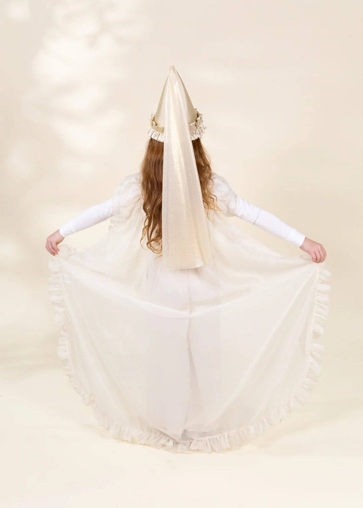 A child wearing a white and cream Spanish Santa Muerte costume, viewed from behind, holding out the wide ruffled skirt with a tall veil and cone headpiece.
