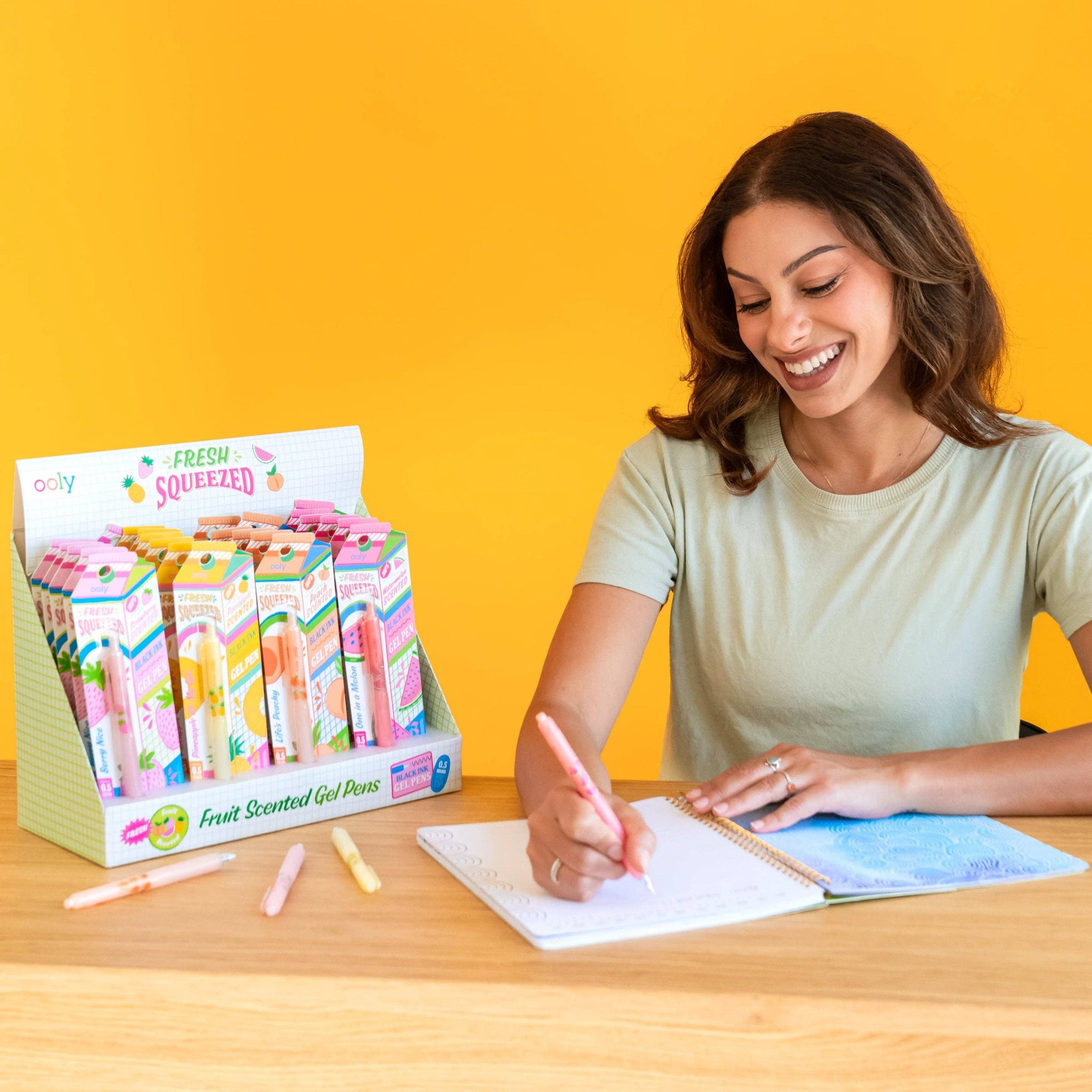 Woman writing with a pink gel pen at a desk with a display of colorful gel pens in the background.