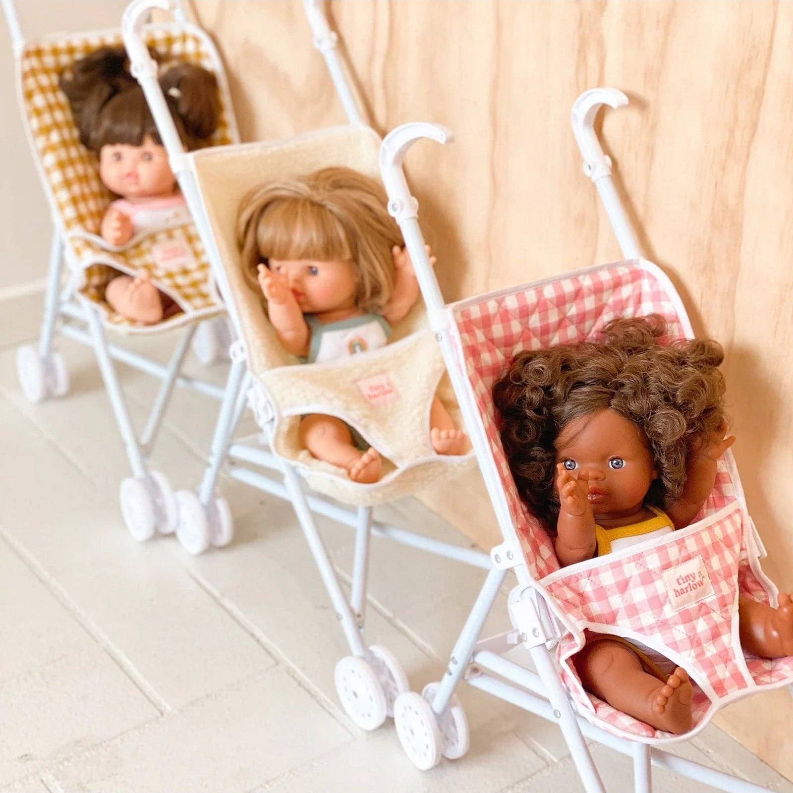 Three dolls in miniature strollers with checkered seats on a light wooden floor.