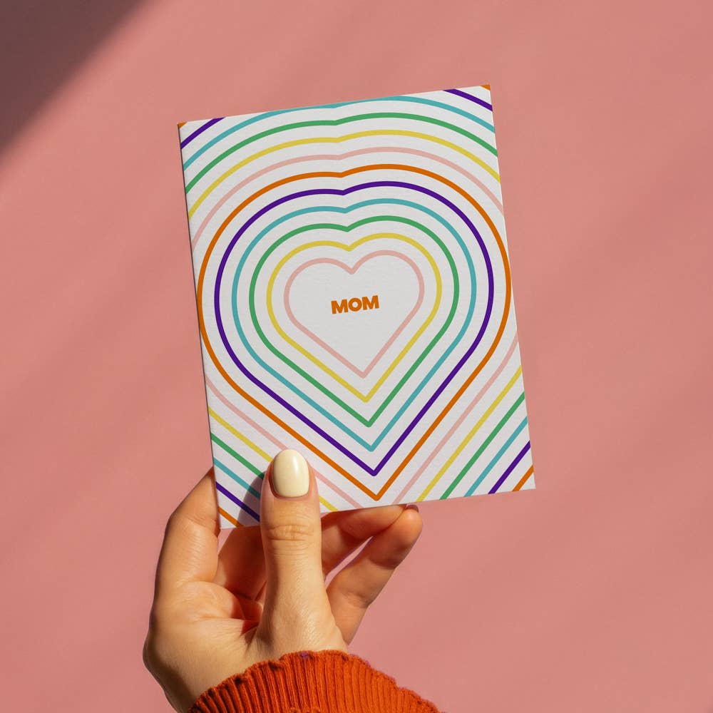 Hand holding a card with rainbow heart design and 'Mom' text on a pink background