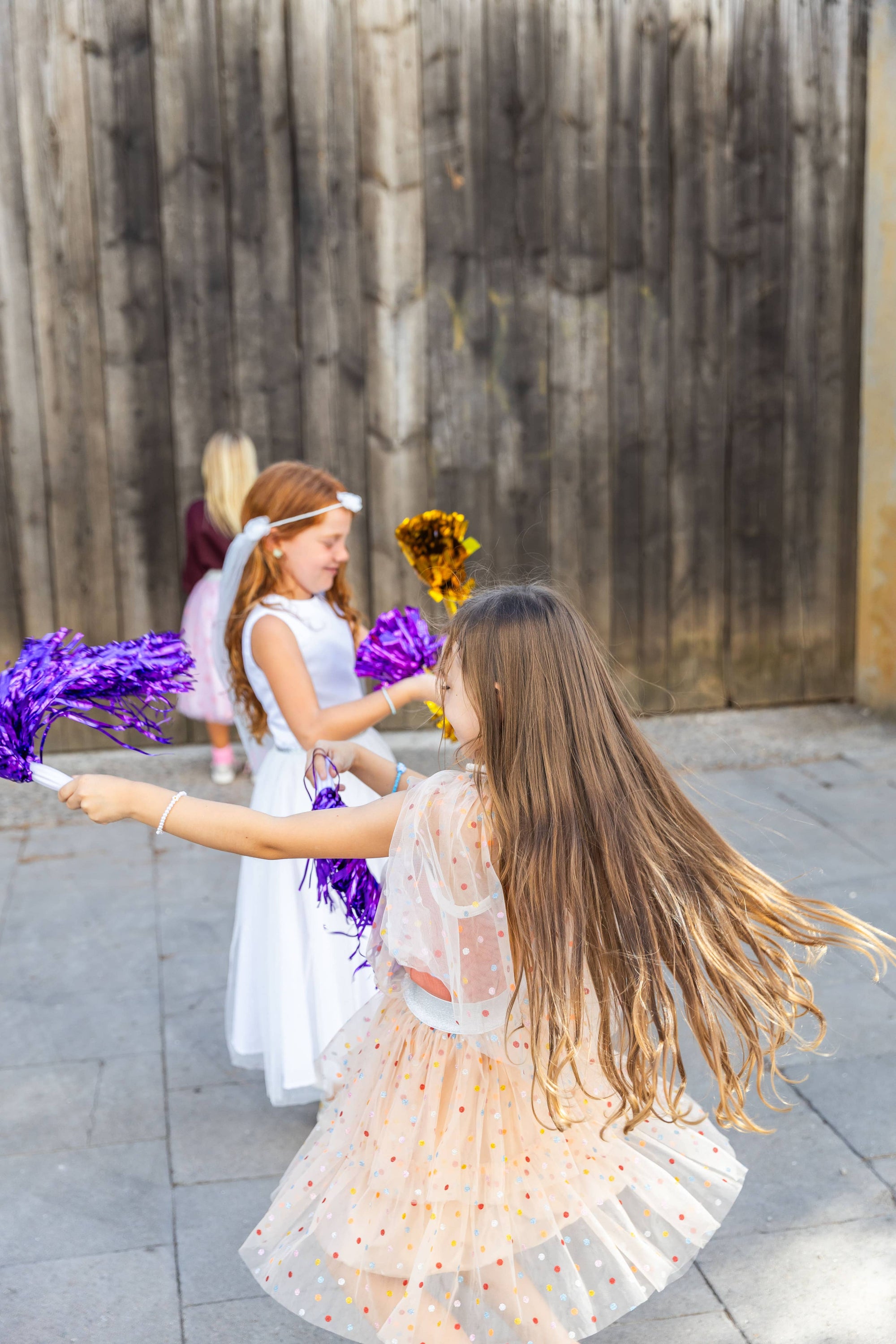 Two young girls in white dresses with purple pom-poms on a stone pavement.