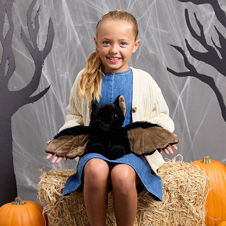 Young girl holding a plush bat in front of a Halloween-themed background with pumpkins and hay.