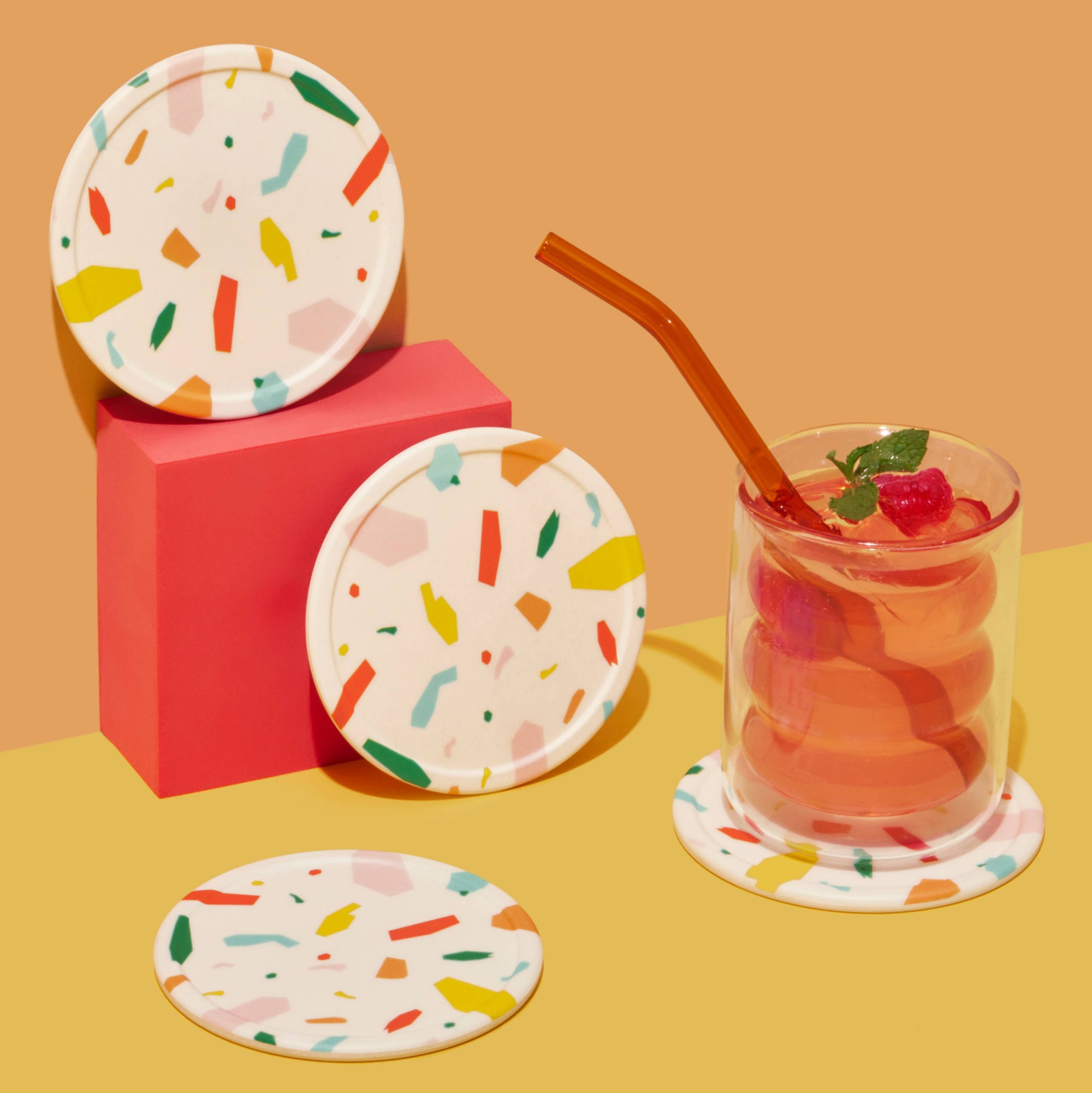 Colorful terrazzo-patterned coasters with a glass of juice on a yellow surface.