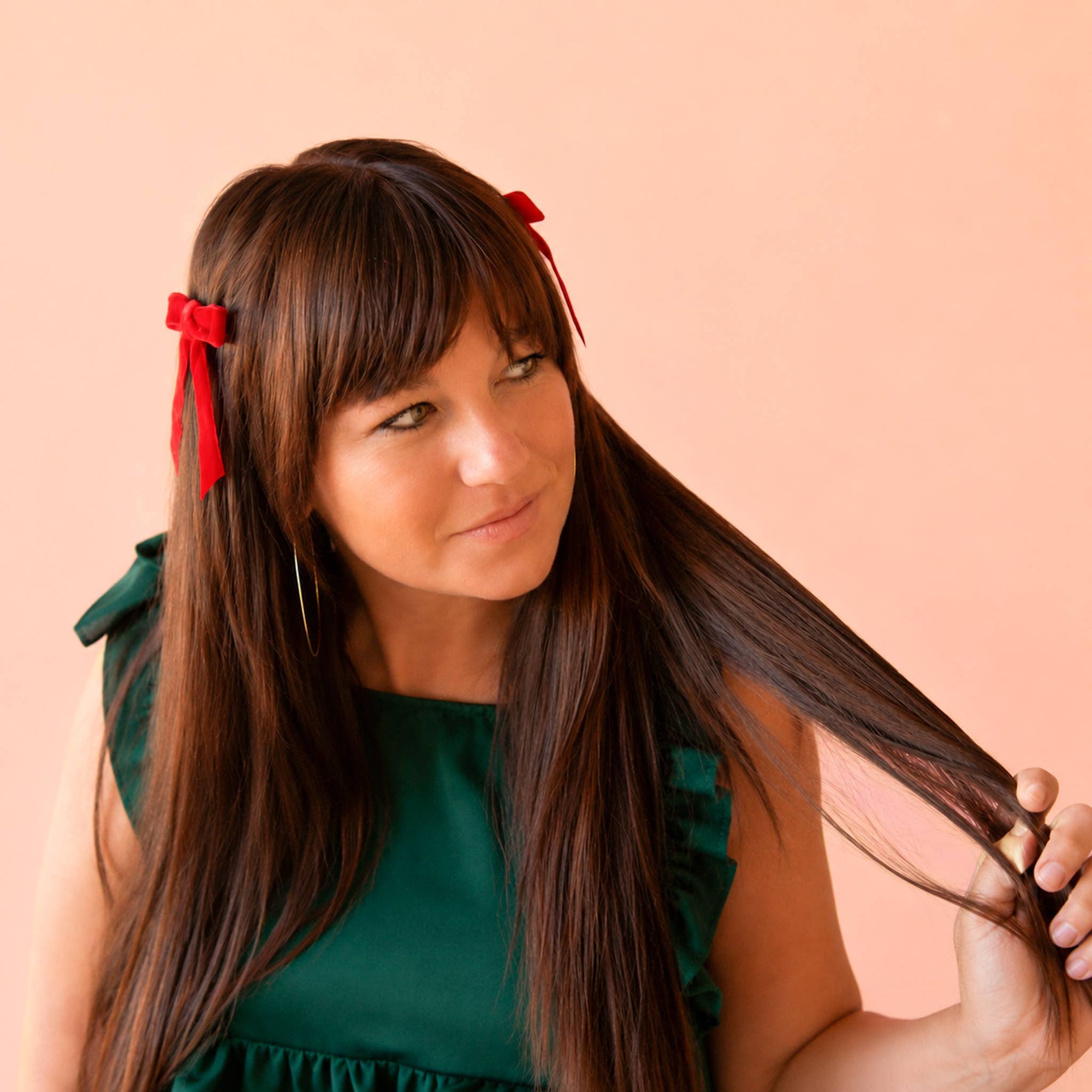 A woman with long, straight brown hair and bangs, wearing a dark green dress and red ribbon hair clips, holding a section of her hair against a solid peach background.