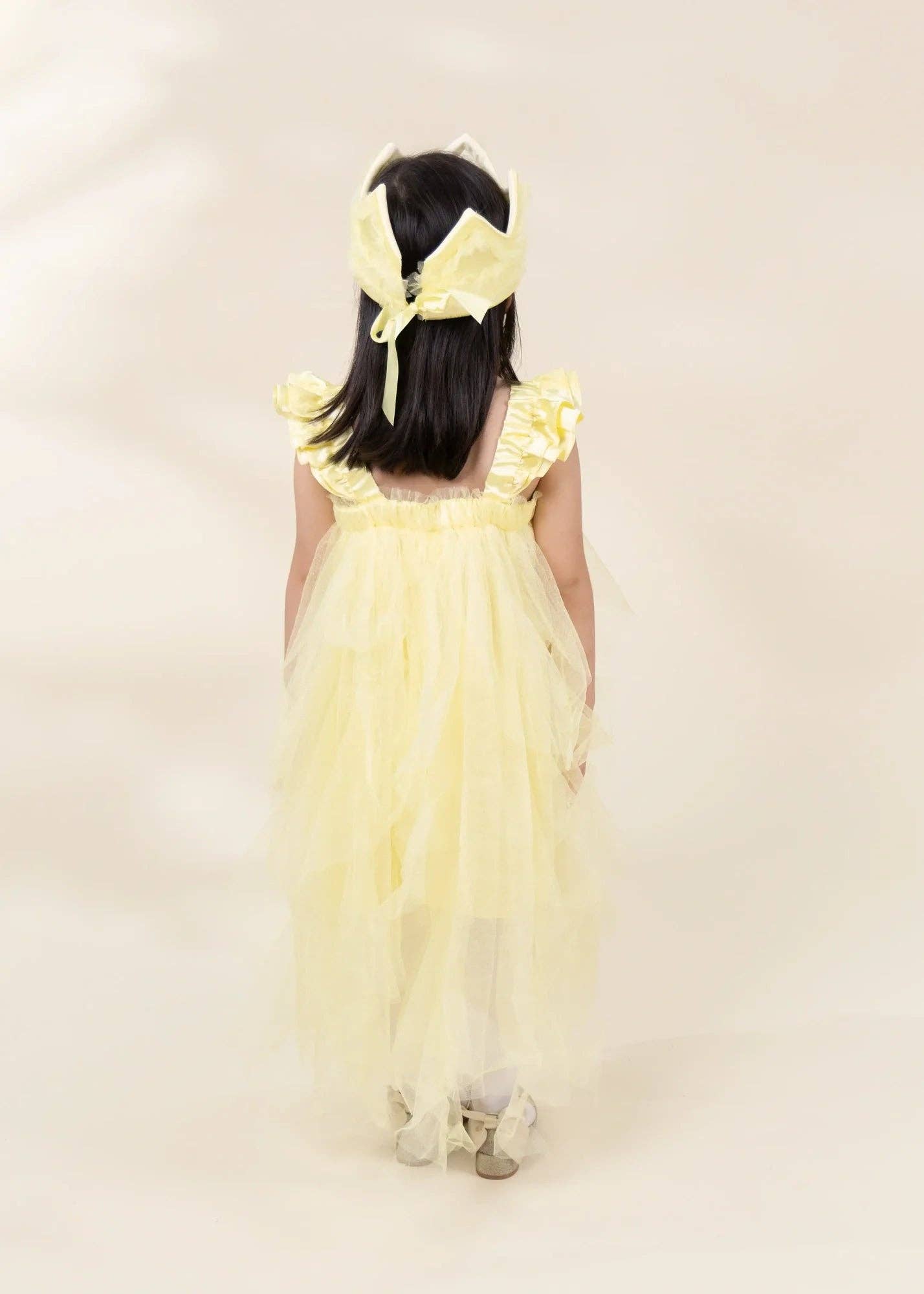 A young girl viewed from behind wearing a pale yellow tulle dress with ruffled shoulder details and a matching bow headband, standing against a plain light beige studio background.