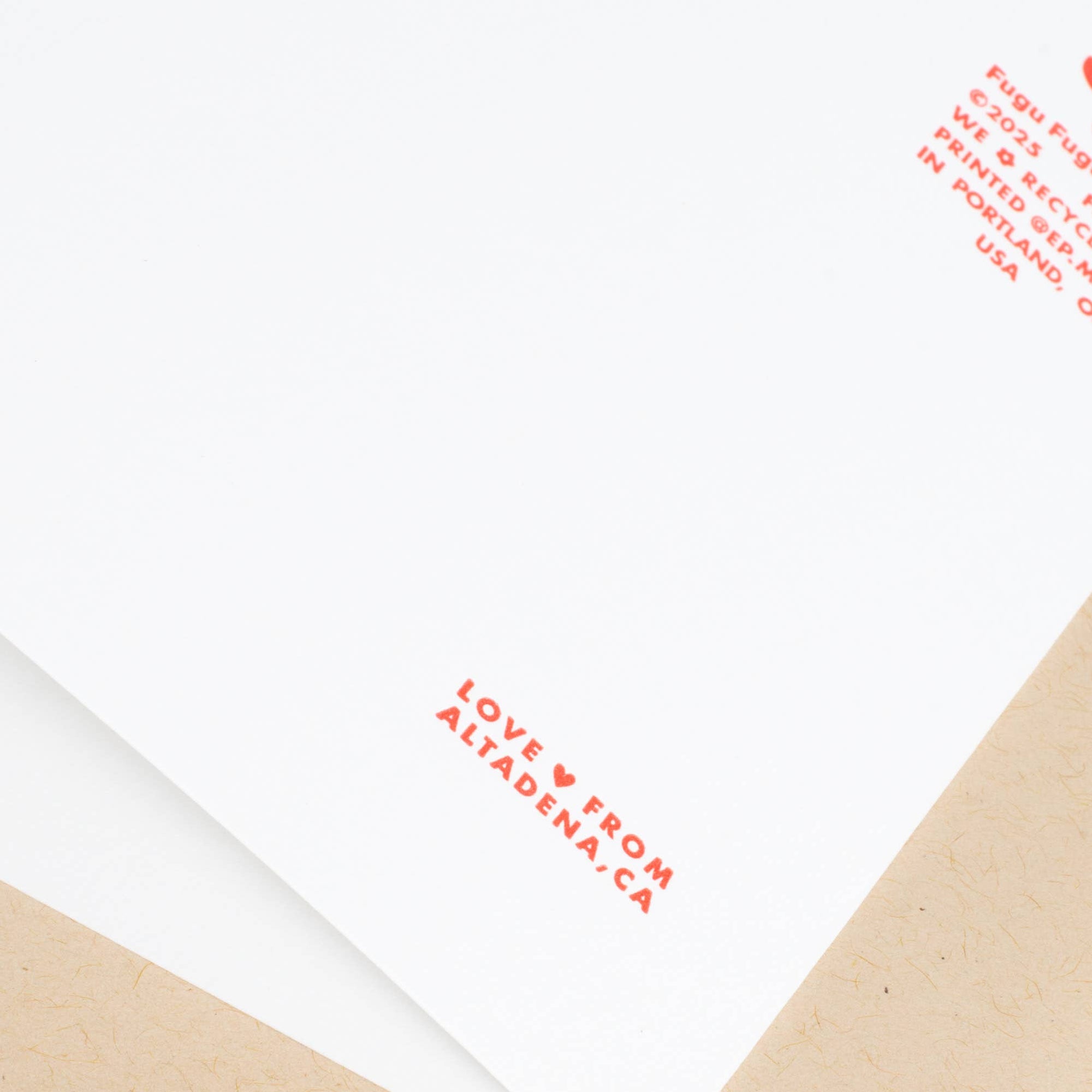 A close-up of a white card or envelope with red printed text reading 'LOVE FROM ALTADENA,CA' and additional red text in the top right corner, layered over a kraft brown paper backing on a white background.