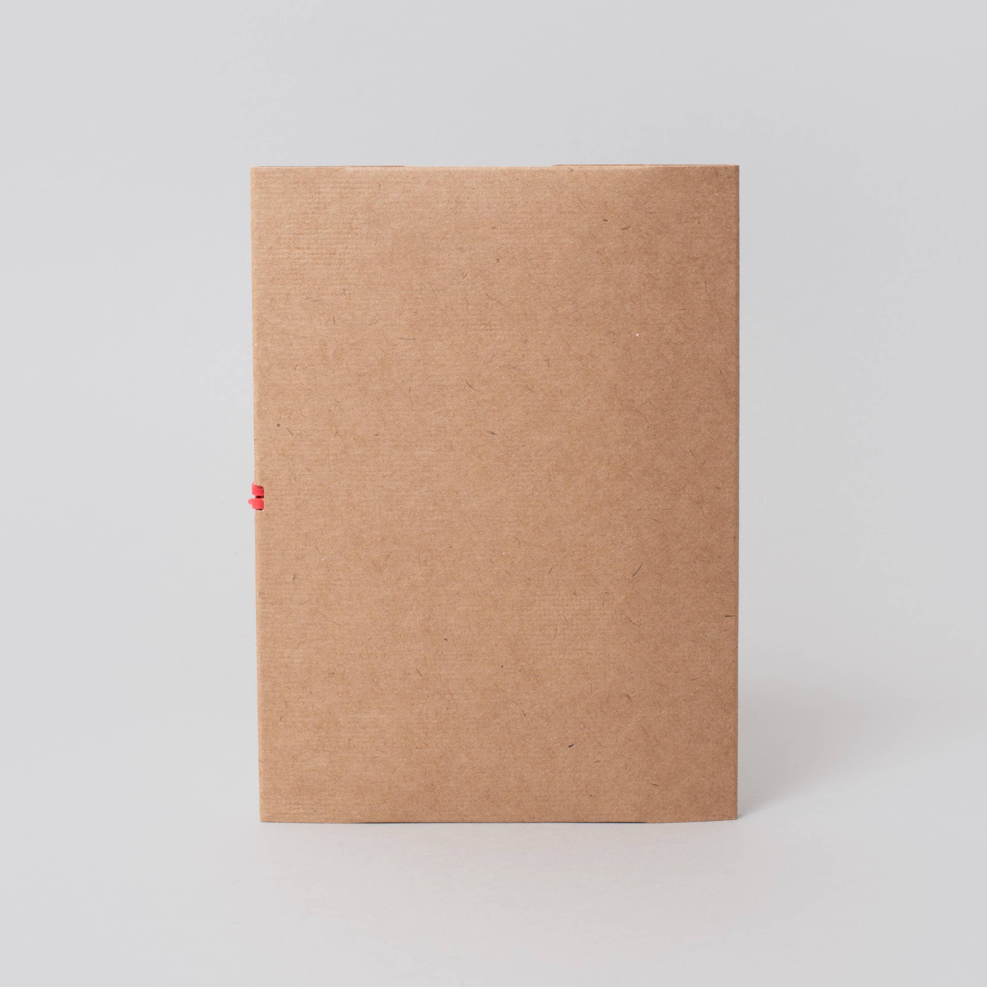 A plain brown kraft paper envelope or notebook cover standing upright on a light gray background, with a tiny red string tied at the left edge.