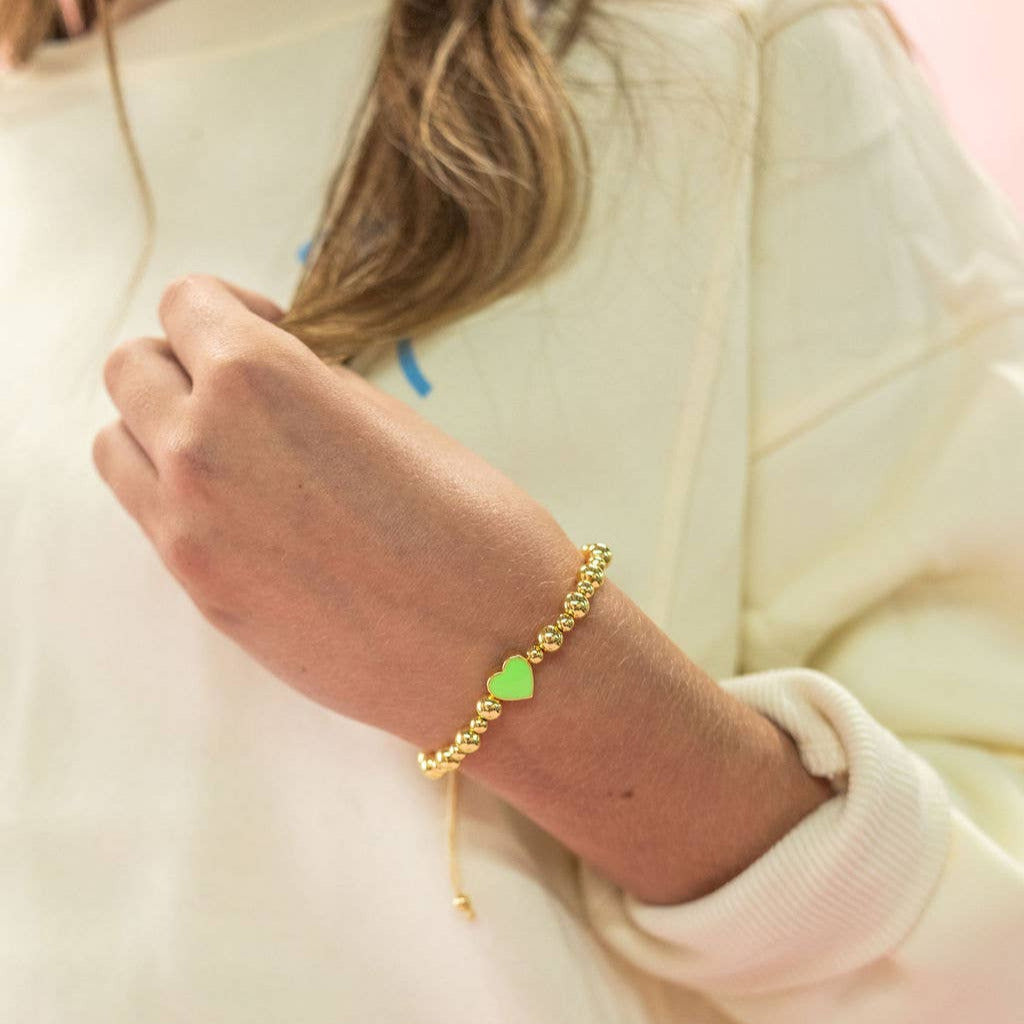 Gold bracelet with a green heart charm on a wrist against a neutral background