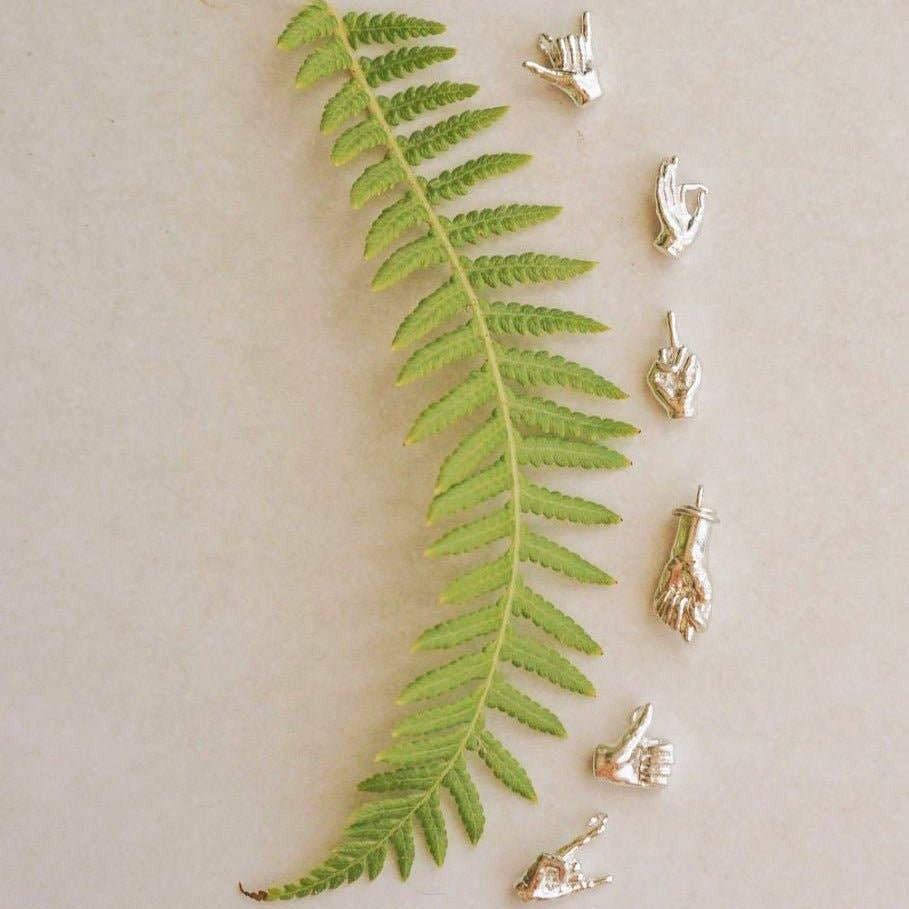 A green fern leaf is laid diagonally on a light beige background, with several small metallic hand-shaped jewelry pieces arranged along the right side.