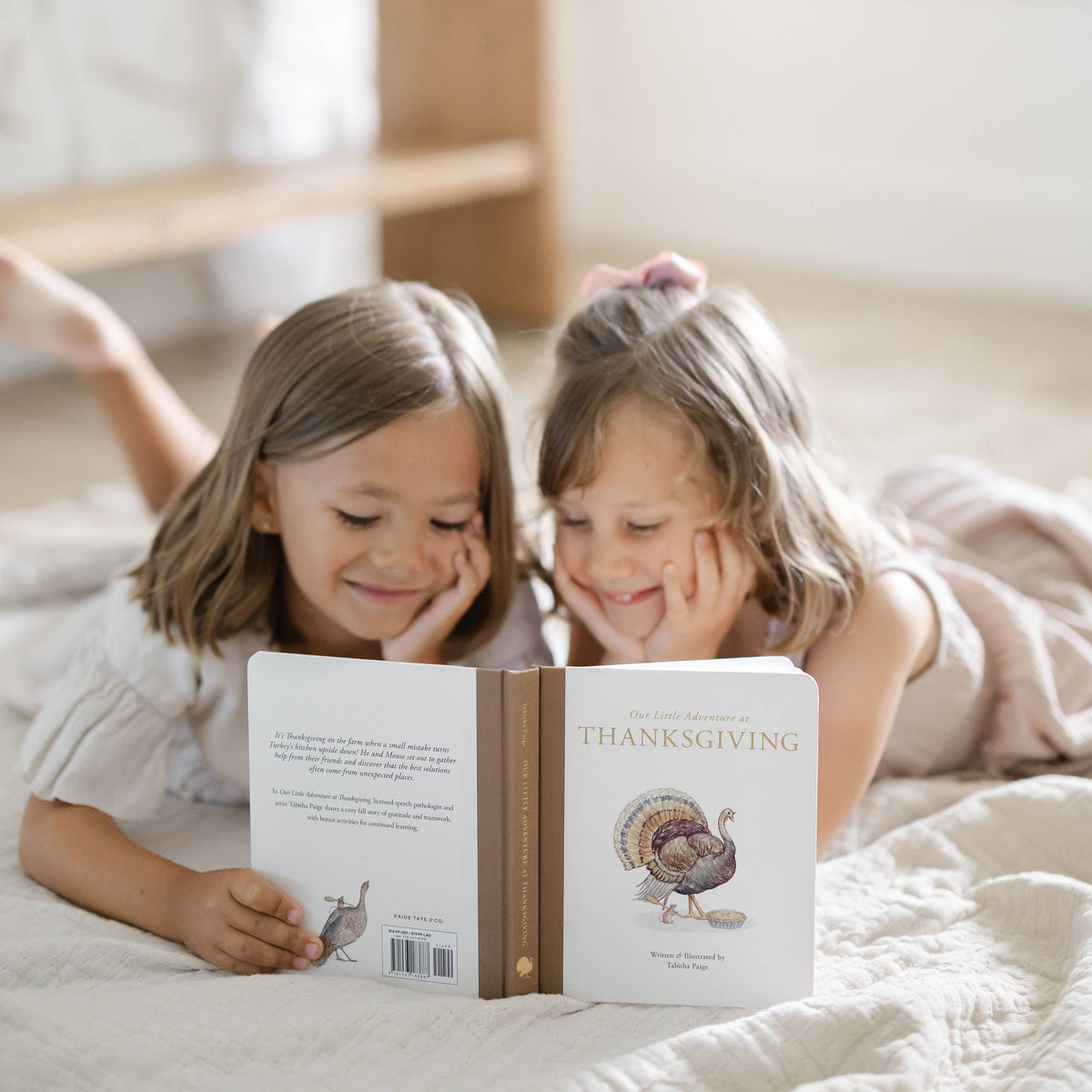 Two young girls lying on a quilted bed, smiling while looking at a personalized children's book titled 'The Little Thanksgiving' with a turkey illustration on the back cover.