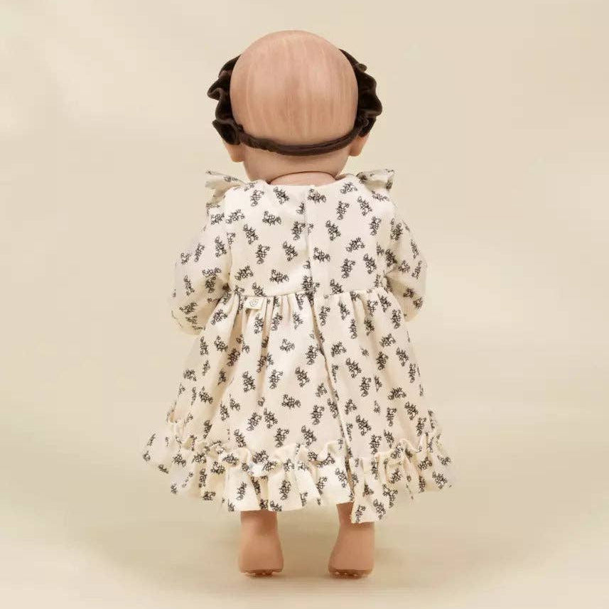 A doll viewed from behind wearing a cream-colored long-sleeve dress with a small black floral pattern and a black headband, set against a solid beige background.