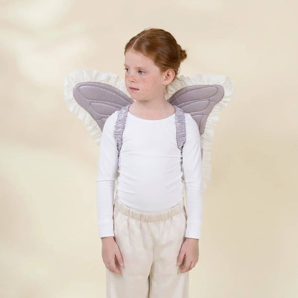 A young girl with red hair tied back stands against a soft cream backdrop wearing a white long-sleeve top, cream pants, and gray angel wings with white ruffled trim.