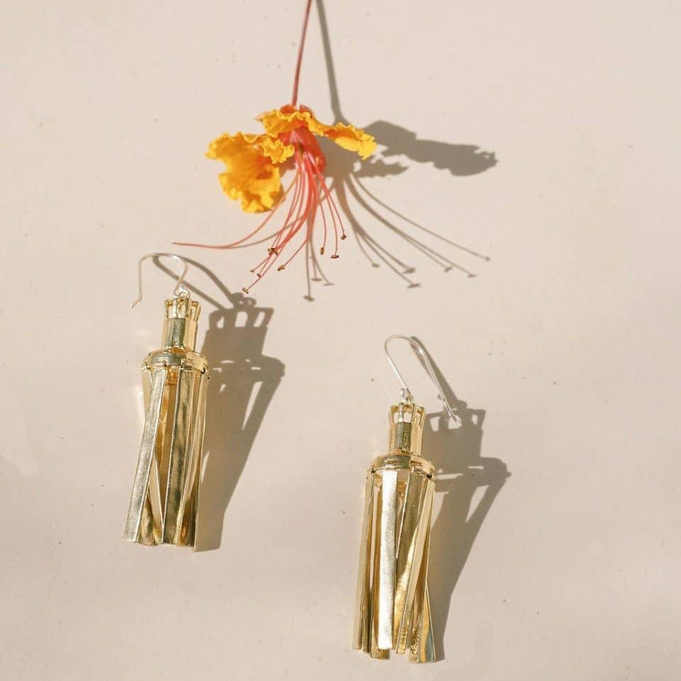 A pair of gold-toned tassel earrings with a twisted cylindrical design, displayed on a light beige background with a yellow flower prop casting shadows.