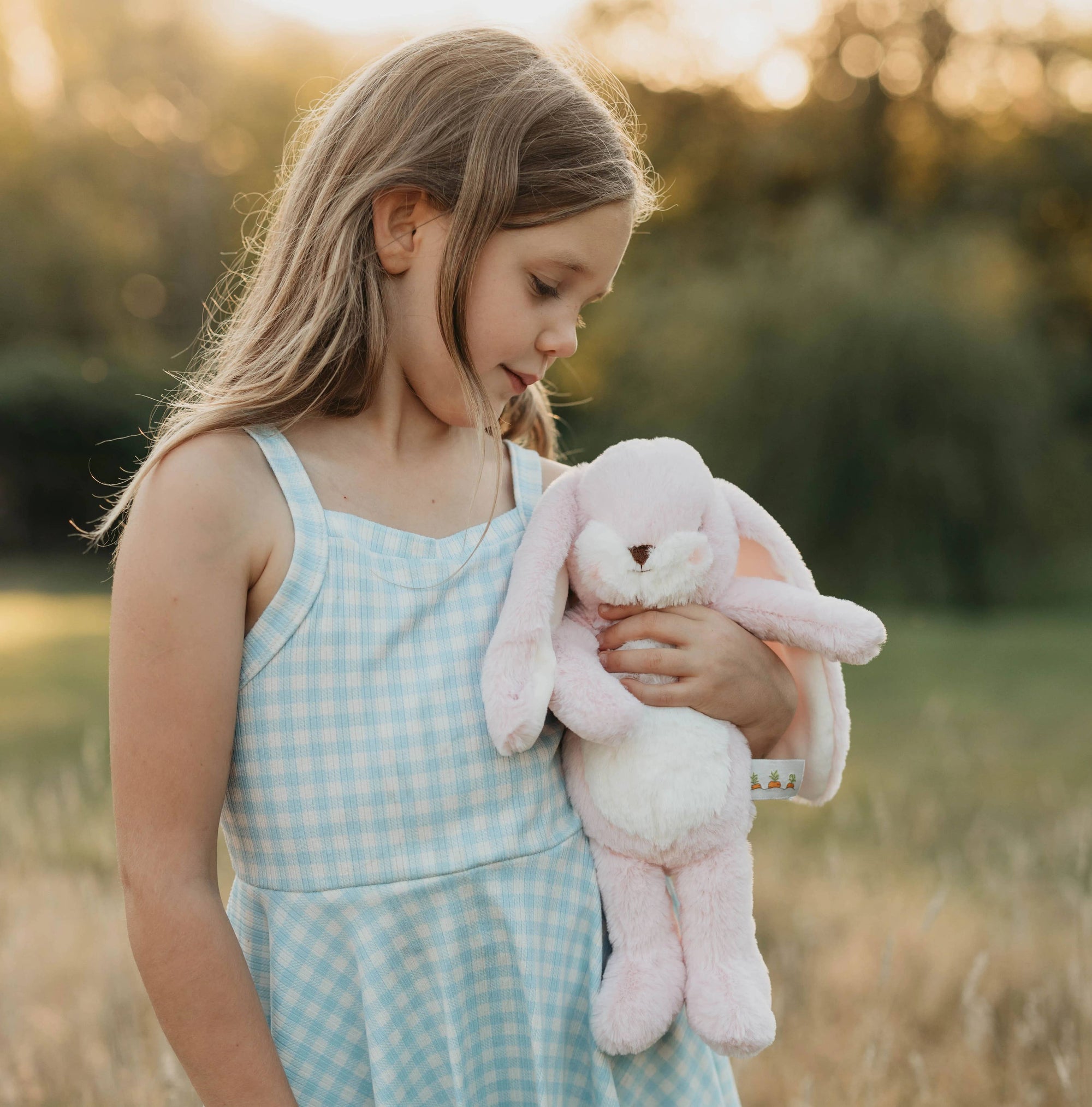 Young girl holding a pink bunny toy in a field during sunset