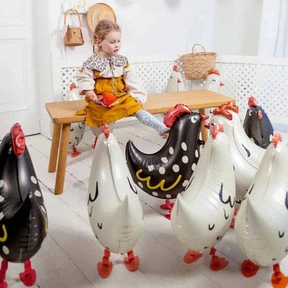 A young girl in a yellow dress sits on a wooden bench in a bright room, surrounded by inflatable farm animal balloons, including several white chickens with red feet and two black-and-white chickens with red combs.