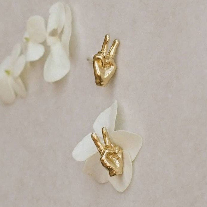 Two small gold hand-shaped earrings, one plain and one nestled among white flower petals, arranged on a light beige background.