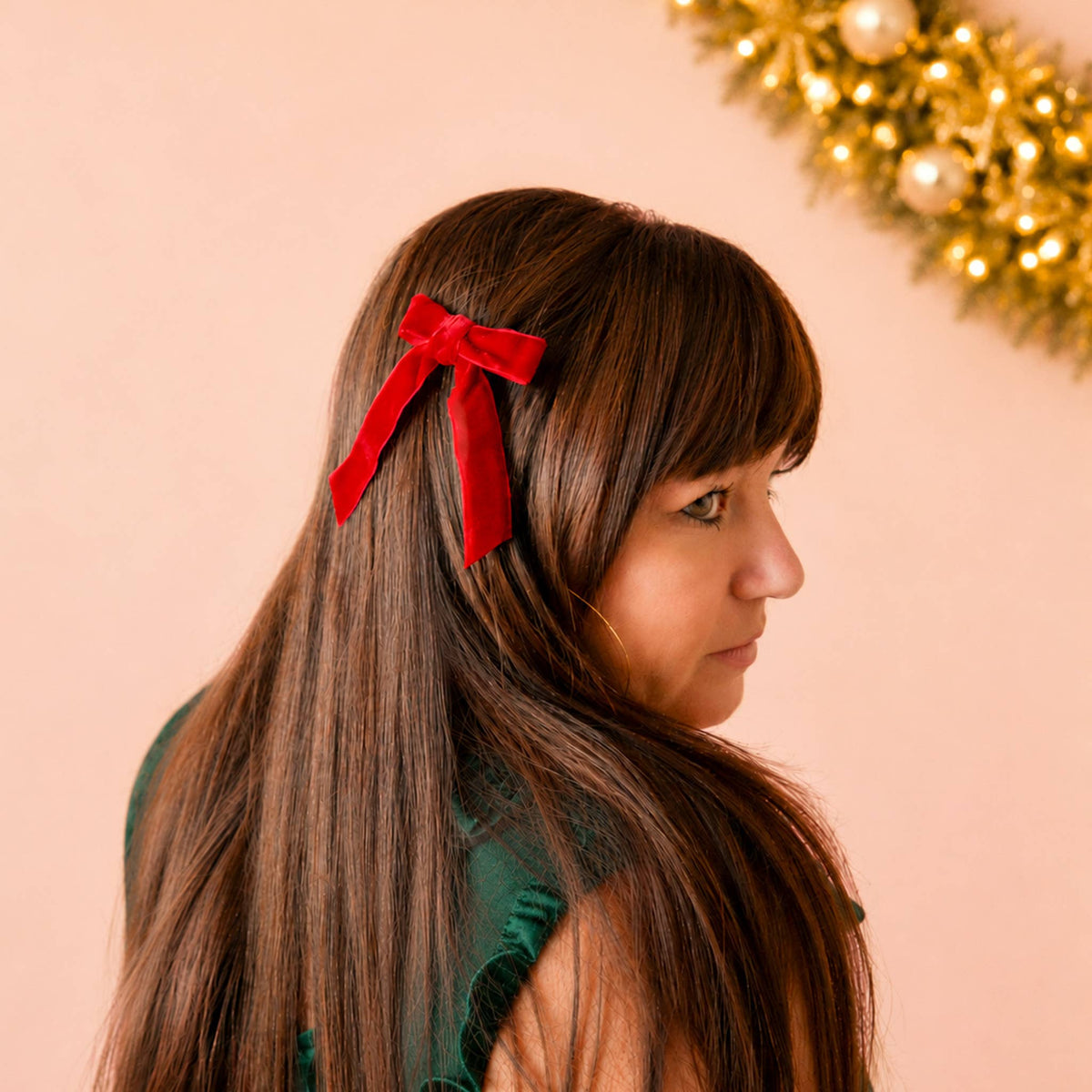 A woman with long brown hair and bangs shown in profile, wearing a bright red bow hair clip. The background is a soft peach-pink color with a warm, glowing gold holiday garland visible in the top right corner.