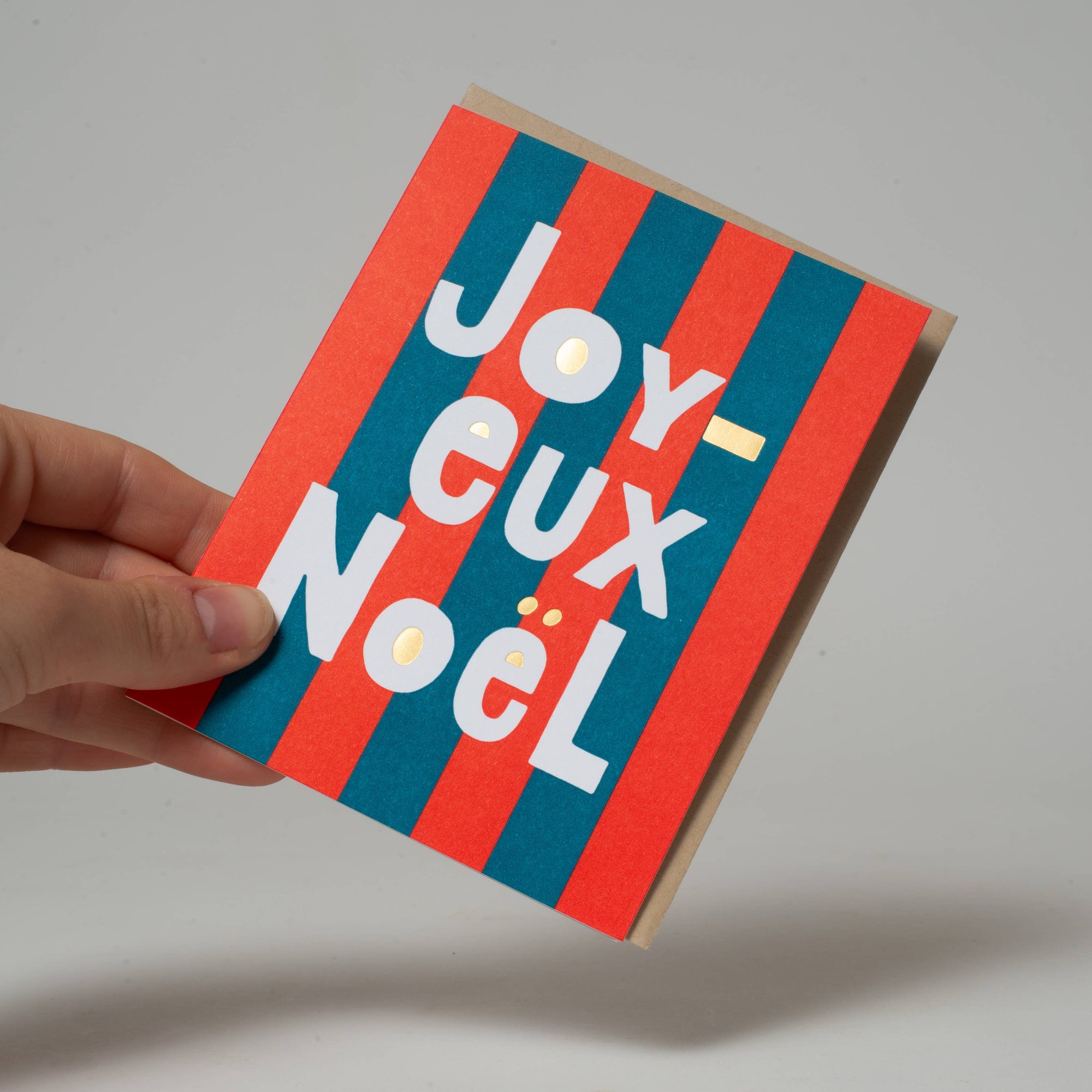 Hand holding a small card with 'Joyeux Noel' text on red and blue striped background