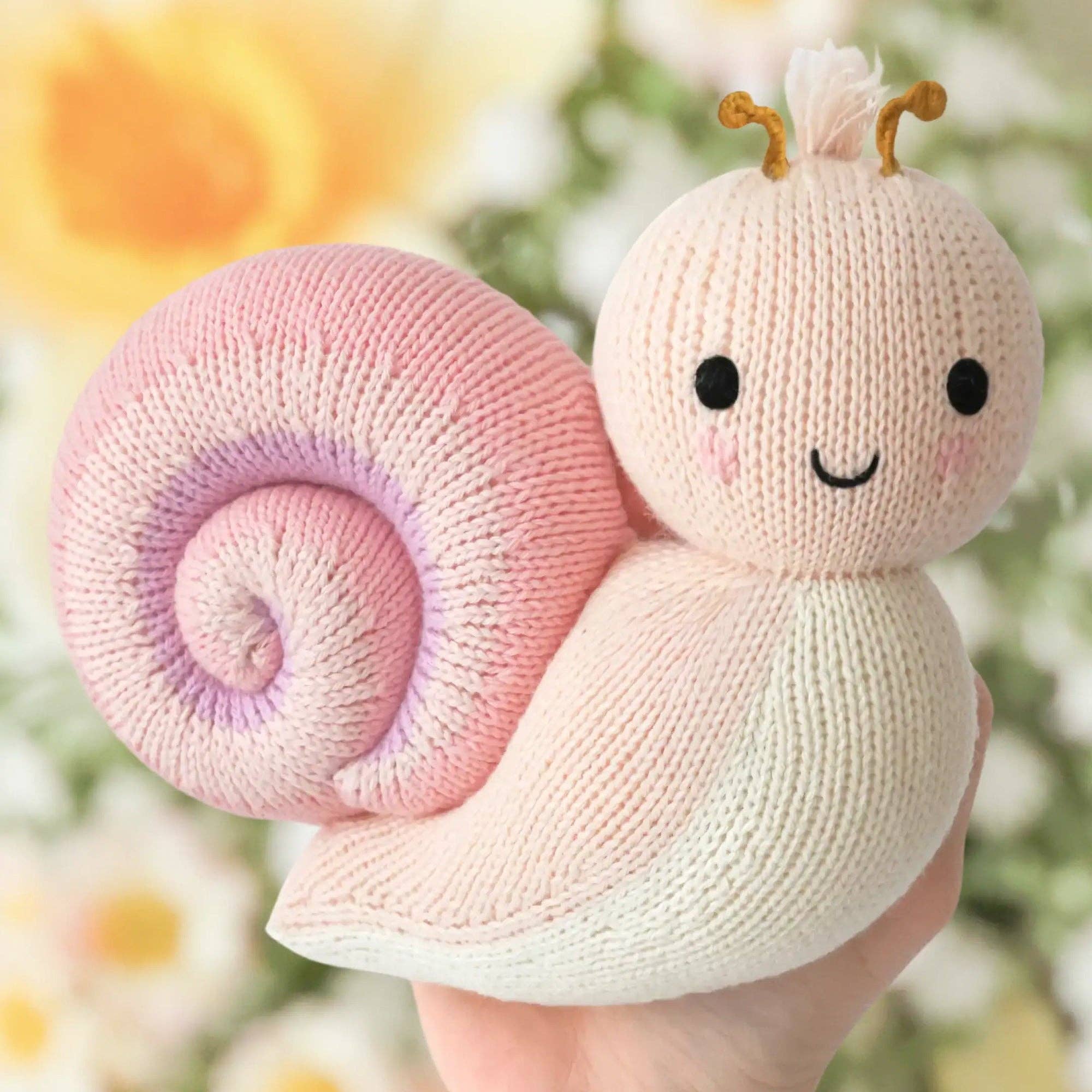 Knitted snail toy held in a hand with a blurred floral background