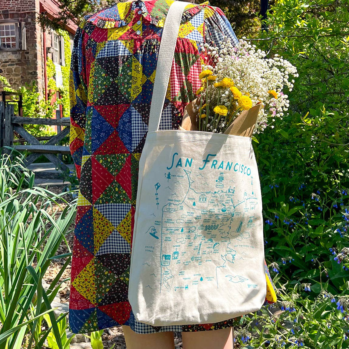 Person holding a colorful quilt and a tote bag with 'San Francisco' printed on it, standing in a garden.