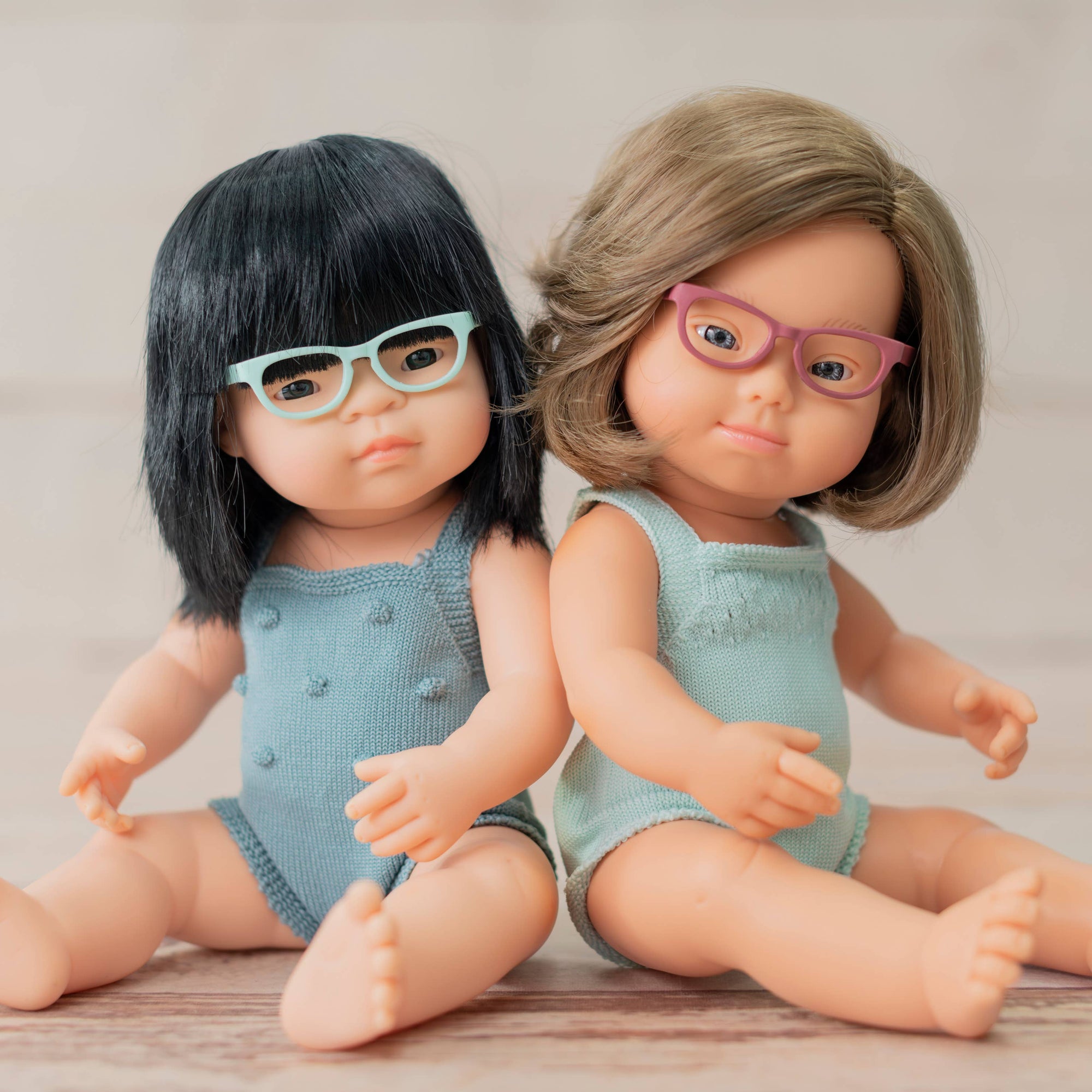 Two dolls with different hairstyles and glasses sitting on a wooden surface.