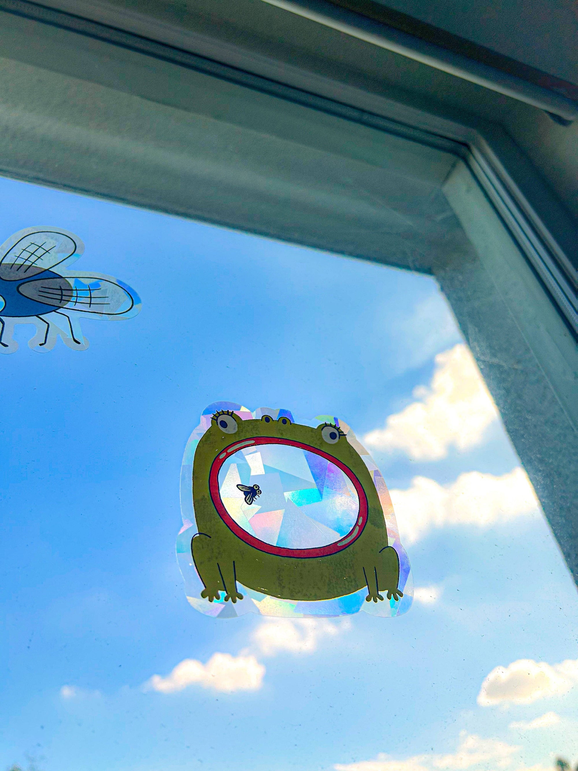 Decorative window sticker of a green frog with a red circle on a clear blue sky background.
