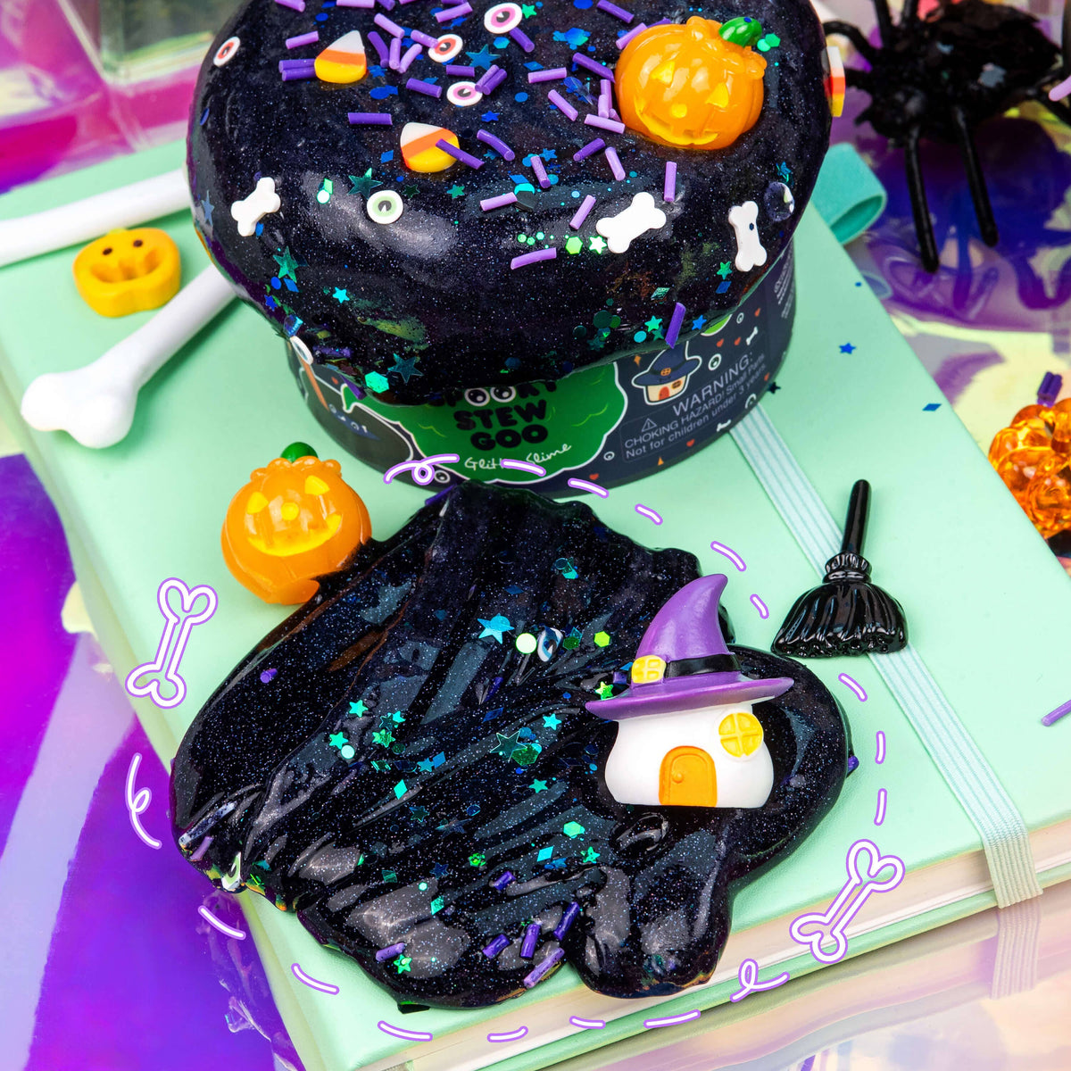 Halloween-themed decorative slime displayed on a mint notebook, with a large black glitter slime mound topped with candy corn, sprinkles, and white bone shapes, plus a smaller black slime puddle shaped like a witch hat house with a purple witch hat. Surrounding props include a broom, bones, jack-o'-lanterns, and a spider decoration.