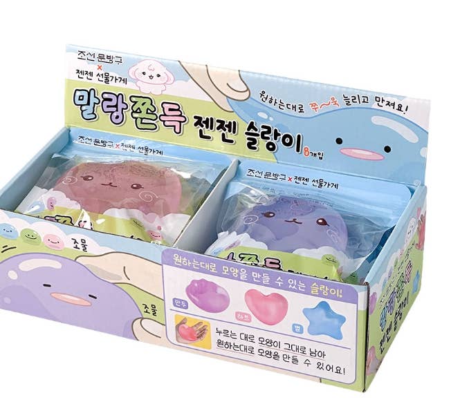 A retail display box containing two small packaged plush toy pieces, with colorful printed packaging in Korean and cartoon illustrations. The left package contains a pink plush, and the right package contains a purple plush.