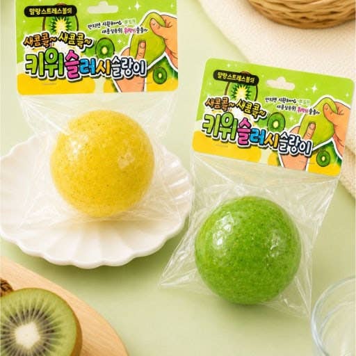 Two packaged round squishy toys, one yellow and one green, displayed on a pale green surface with fruit props and a white plate. The products are in clear plastic bags with bright green and yellow retail cards featuring Korean text.