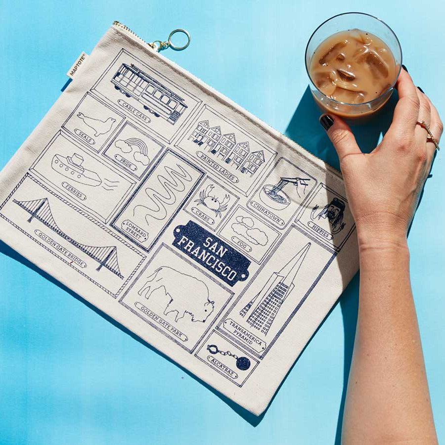 A cream canvas zip pouch printed with San Francisco landmarks in navy blue, held by a hand on a bright blue background next to a glass of iced coffee.