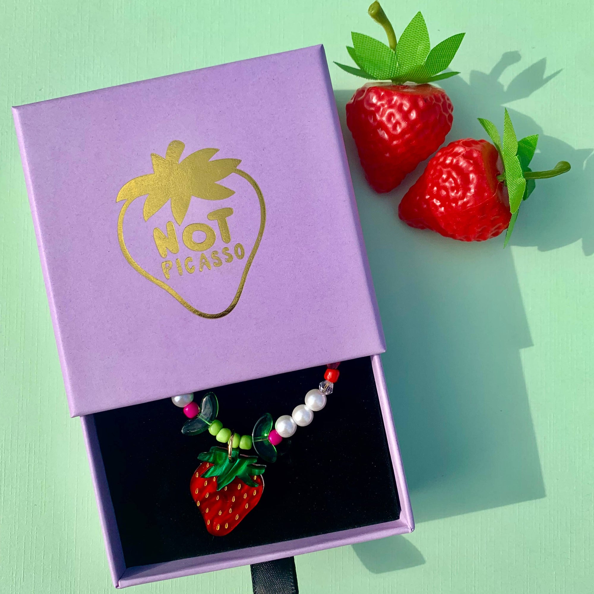 Strawberry-themed necklace in a 'Hot Tomato' branded box with two strawberries on a light green background