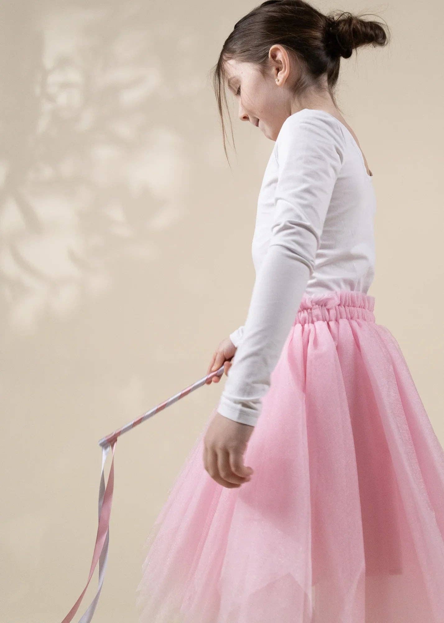 A young girl in a white long-sleeve top and a fluffy pink tulle skirt holding a pink and white striped ribbon, shown from a side-back angle against a warm beige background with soft leaf shadows.