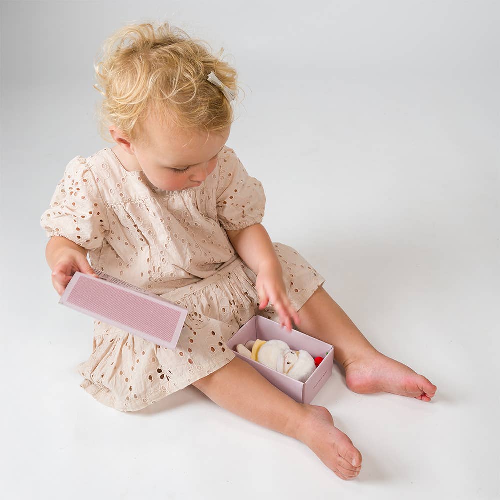 Child in a light dress sitting on a white surface with a small box containing toys.