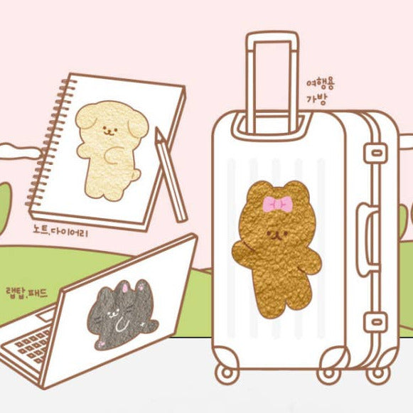 A cute travel-themed illustration showing a white suitcase with a teddy bear sticker, a notebook with a dog doodle, and a laptop with a cat doodle, all on a soft pink and green background.