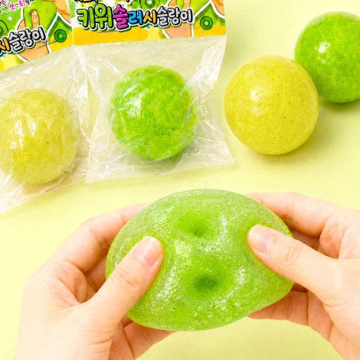 A person's hands are shown squeezing a green squishy toy, with several similar green and yellow-green squishies and their plastic packaging visible on a pale yellow background.
