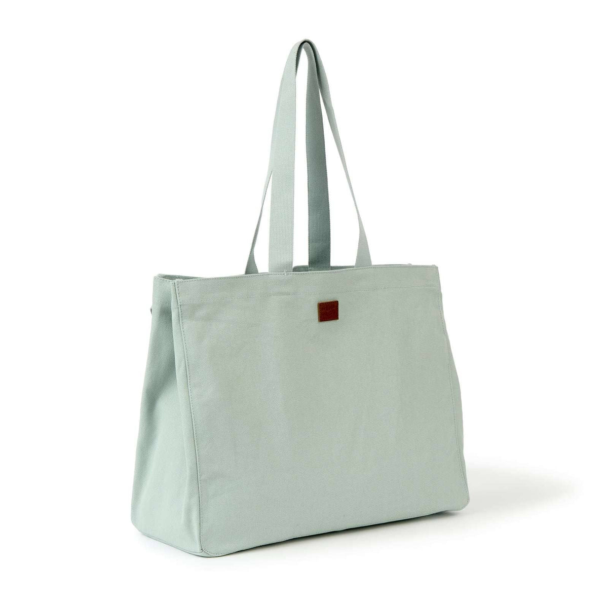 A sage green canvas tote bag with dual shoulder straps and a small dark red square patch on the front, shown on a clean white background.