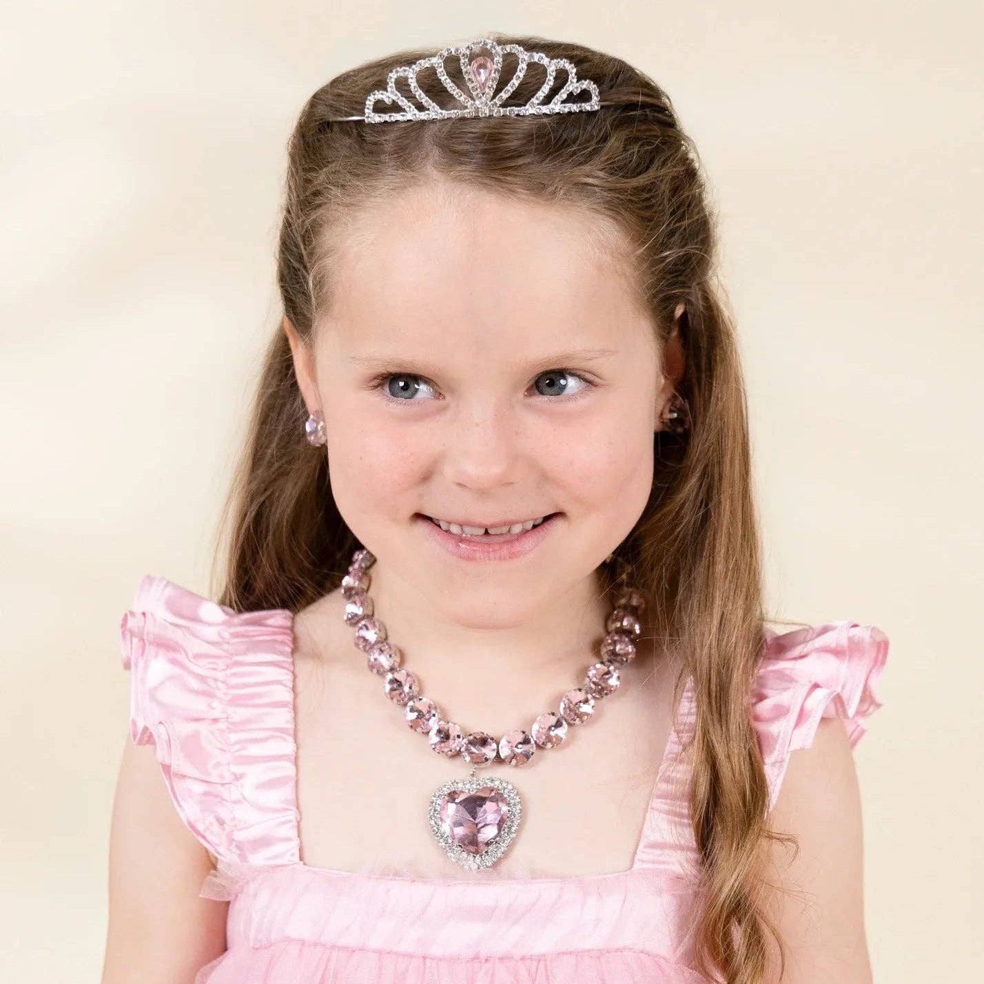 A young girl with long light brown hair wearing a pink dress, a silver tiara, a pink beaded necklace, and matching earrings, set against a plain beige background.