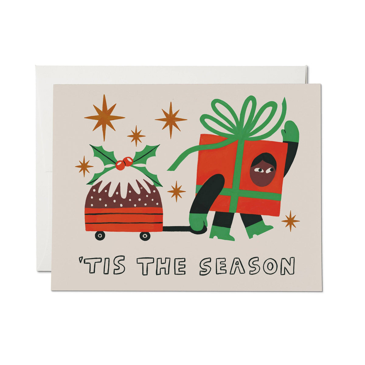 A Christmas greeting card with a beige background, featuring a stylized illustration of a pudding in a sleigh pulled by a gift box with a person peeking out, plus the text “'TIS THE SEASON” at the bottom. A white envelope is placed behind the card.