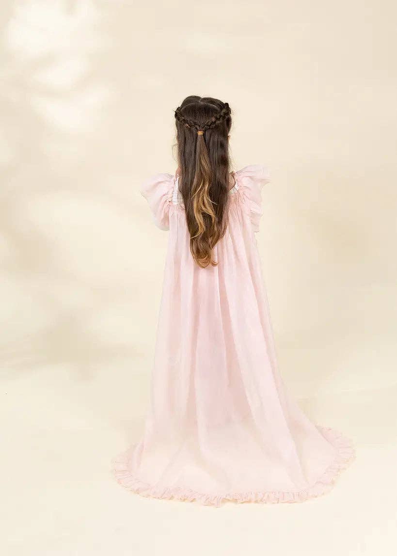 A young girl viewed from behind wearing a long, flowing light pink dress with ruffled sleeves and a long train, standing against a soft cream background.