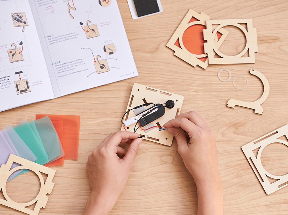 Hands assembling a wooden craft project on a table with a manual and color swatches.