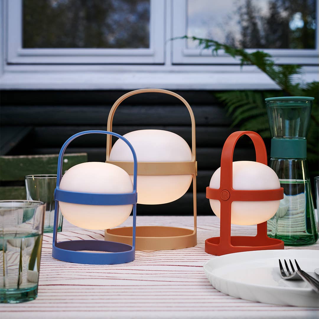 Three portable lamps in blue, beige, and red on a table with glasses and cutlery.