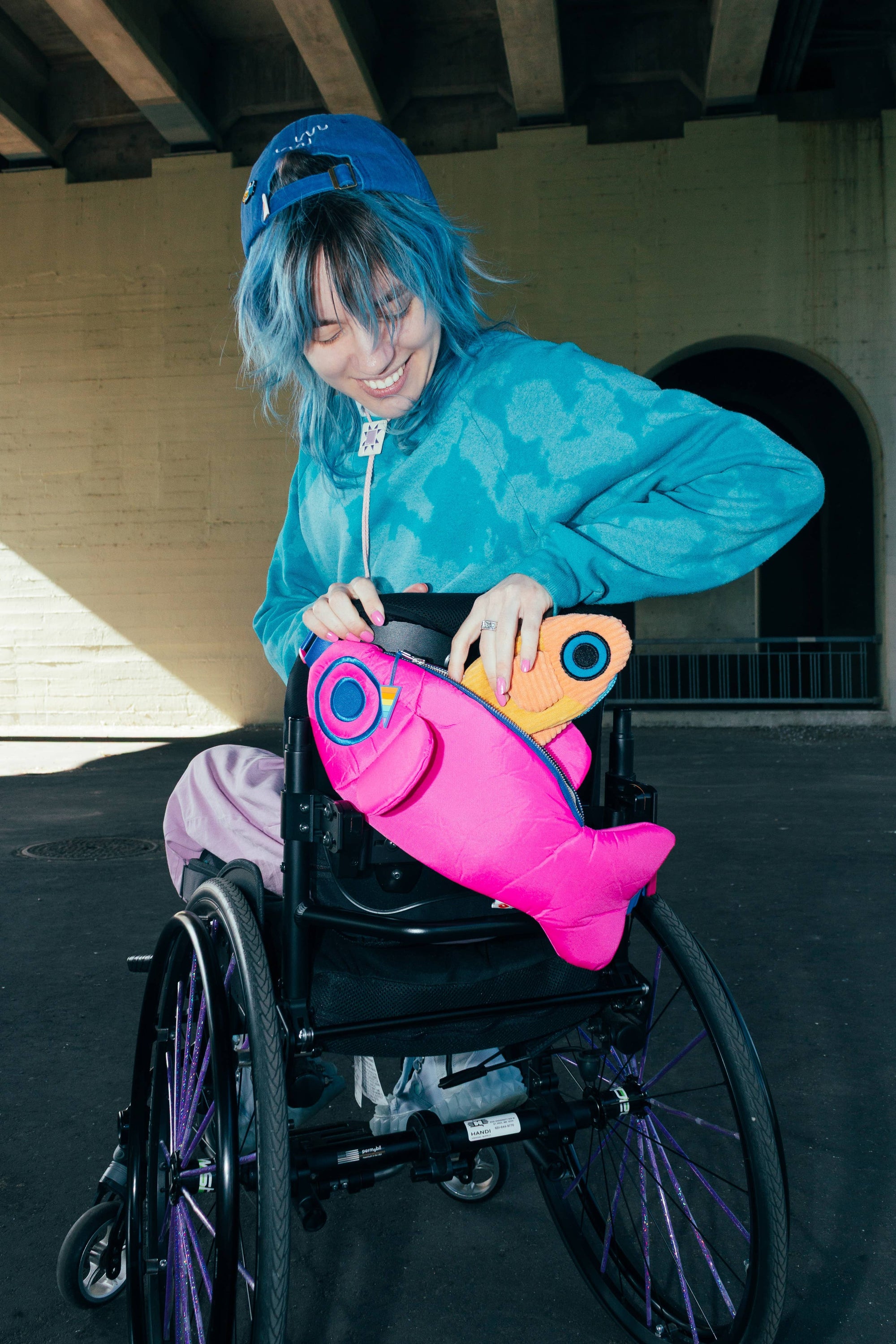 Person with blue hair in a wheelchair holding a colorful fish-shaped bag.